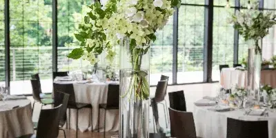 A tall, elegant floral arrangement with white flowers and greenery in a clear glass vase, placed on a set wedding reception table.