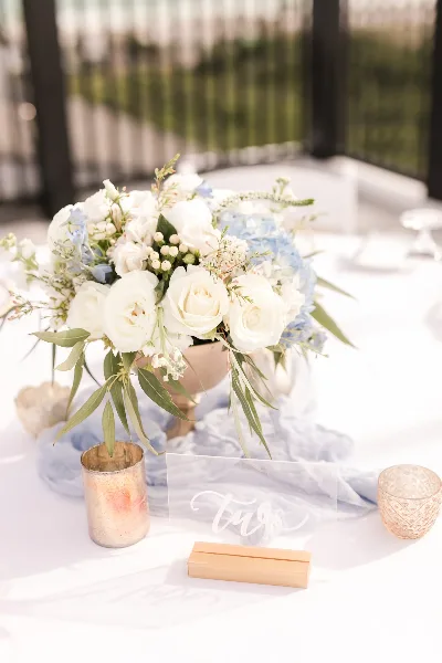 A wedding table setting featuring a white and blue floral arrangement in a vase, accompanied by candles and a table number on a white tablecloth.