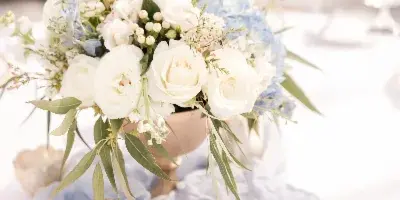A wedding table setting featuring a white and blue floral arrangement in a vase, accompanied by candles and a table number on a white tablecloth.