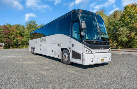 A white Kingz Transportation coach bus parked in a gravel lot with trees in the background.