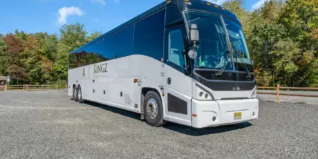 A white Kingz Transportation coach bus parked in a gravel lot with trees in the background.