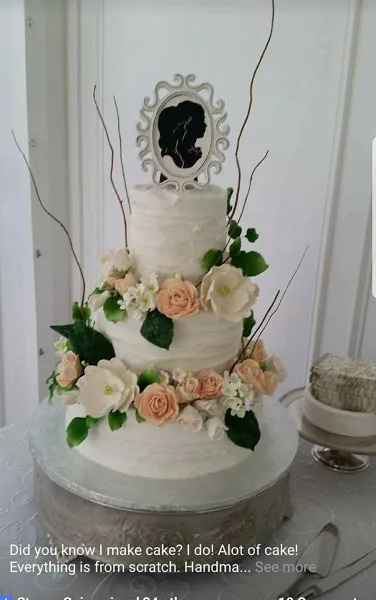 A three-tiered white wedding cake decorated with cream and peach roses, greenery, and a framed silhouette topper.