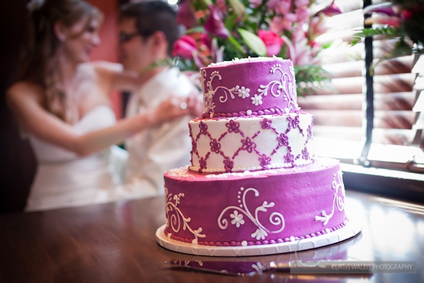 A three-tiered purple and white wedding cake with floral decorations on a table, with a bride and groom in the background.