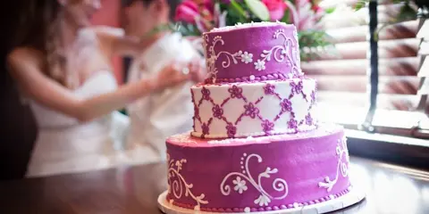 A three-tiered purple and white wedding cake with floral decorations on a table, with a bride and groom in the background.