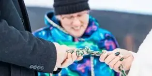 A close-up of a handfasting ceremony where a couple's hands are tied with a rope by an officiant in a snowy outdoor setting.