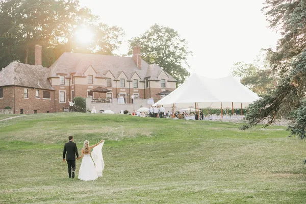 A bride and groom walking across a grassy lawn toward a large white event tent, with the historic brick manor house in the background.