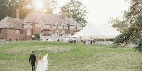 A bride and groom walking across a grassy lawn toward a large white event tent, with the historic brick manor house in the background.