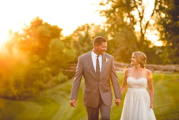 A bride and groom holding hands and walking on a grassy path during golden hour.