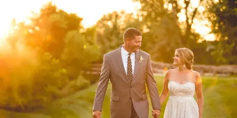 A bride and groom holding hands and walking on a grassy path during golden hour.