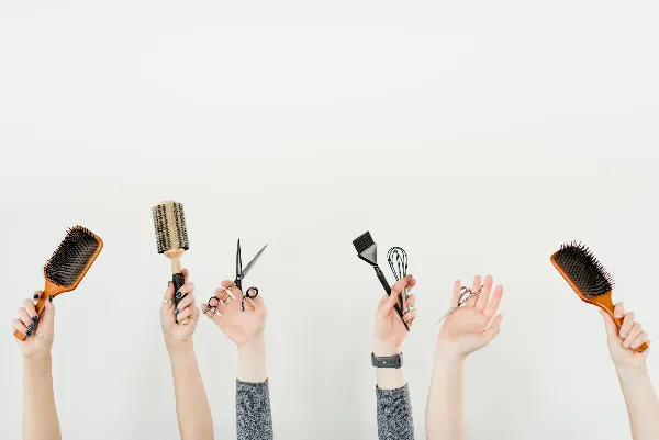 Several hands holding salon tools including scissors, hairbrushes, and a color brush against a white background