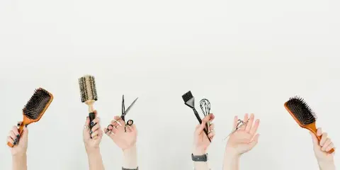 Several hands holding salon tools including scissors, hairbrushes, and a color brush against a white background