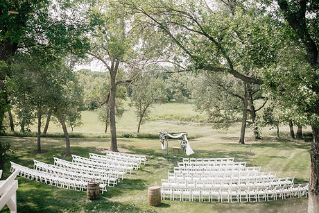 An outdoor wedding ceremony setup featuring rows of white chairs and a decorated altar under trees.