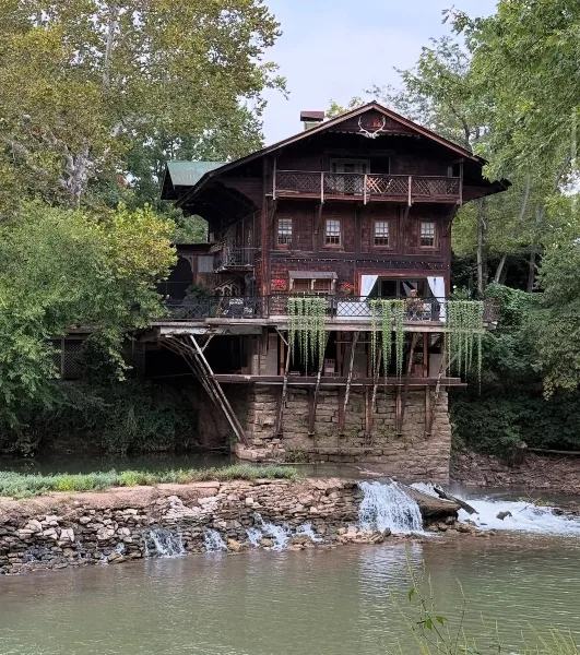 A historic three-story wooden mill building with a stone foundation and waterfall, situated on a river surrounded by trees.