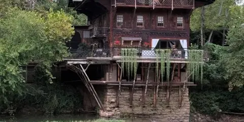 A historic three-story wooden mill building with a stone foundation and waterfall, situated on a river surrounded by trees.