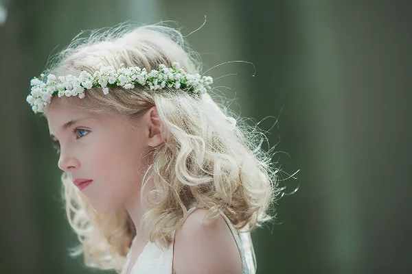 A profile portrait of a young girl with blonde hair wearing a white flower crown, set against a blurred green background.