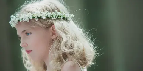 A profile portrait of a young girl with blonde hair wearing a white flower crown, set against a blurred green background.