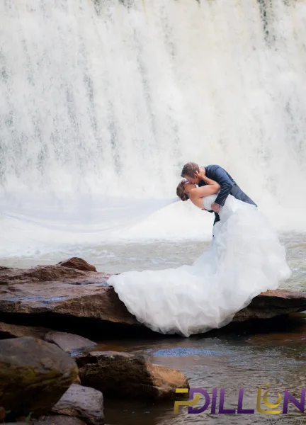 A bride and groom share a kiss on a rock ledge in front of a large waterfall, with the bride's veil and dress flowing in the wind.