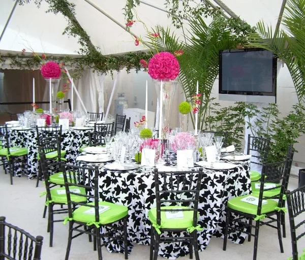 A party setup featuring black chiavari chairs with green cushions, tables with black and white linens, and floral centerpieces under a white tent.