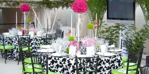 A party setup featuring black chiavari chairs with green cushions, tables with black and white linens, and floral centerpieces under a white tent.