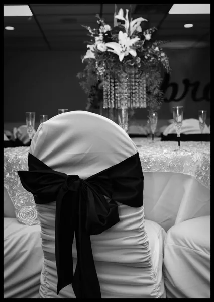 A black and white photograph of a formal event table setting featuring a floral centerpiece and a chair with a black ribbon bow.