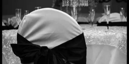 A black and white photograph of a formal event table setting featuring a floral centerpiece and a chair with a black ribbon bow.