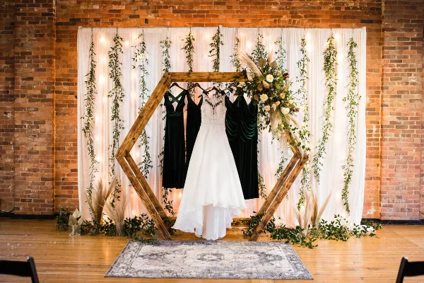 A wedding ceremony setup featuring a wooden hexagonal arch decorated with flowers and greenery, set against a white fabric backdrop with hanging vines and string lights.