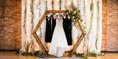 A wedding ceremony setup featuring a wooden hexagonal arch decorated with flowers and greenery, set against a white fabric backdrop with hanging vines and string lights.