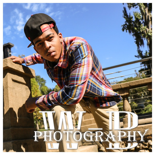 A portrait of a young man wearing a plaid shirt and a black baseball cap, posing outdoors with a stone structure in the background.