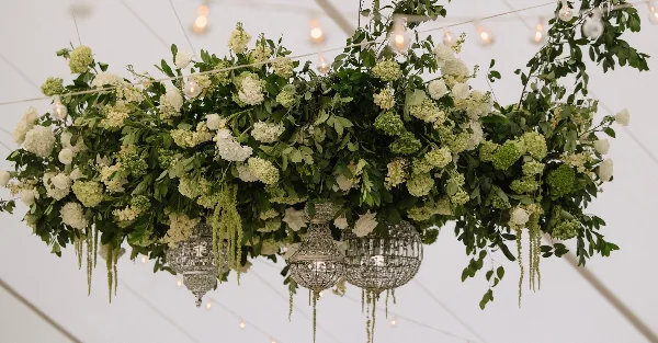 A large, suspended floral installation featuring white roses, hydrangeas, and greenery arranged above ornate silver lanterns.