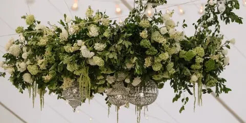 A large, suspended floral installation featuring white roses, hydrangeas, and greenery arranged above ornate silver lanterns.
