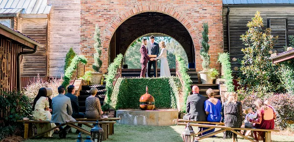 A wedding ceremony in progress under a large brick archway with a couple at the altar and guests seated on benches.