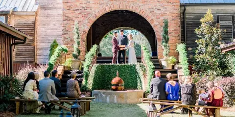 A wedding ceremony in progress under a large brick archway with a couple at the altar and guests seated on benches.