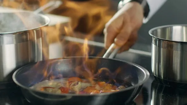 A chef's hands using tongs to cook food in a pan with visible flames in a professional kitchen setting.