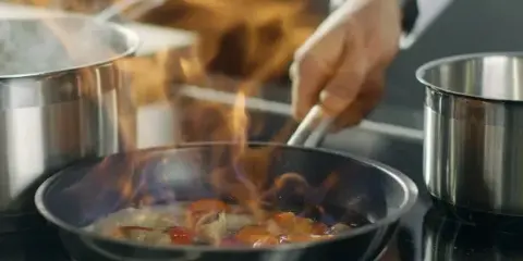 A chef's hands using tongs to cook food in a pan with visible flames in a professional kitchen setting.