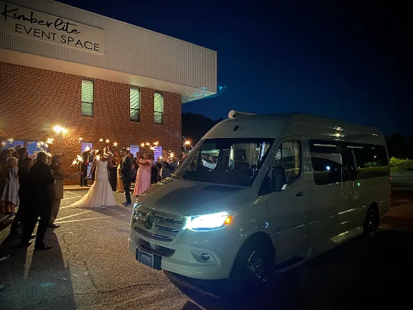 A white Mercedes Sprinter van parked outside a brick event space at night, with a bride and guests holding sparklers.