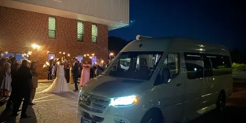 A white Mercedes Sprinter van parked outside a brick event space at night, with a bride and guests holding sparklers.
