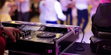 A DJ's hands operating a turntable and mixer at a live event with a crowd dancing in the background.