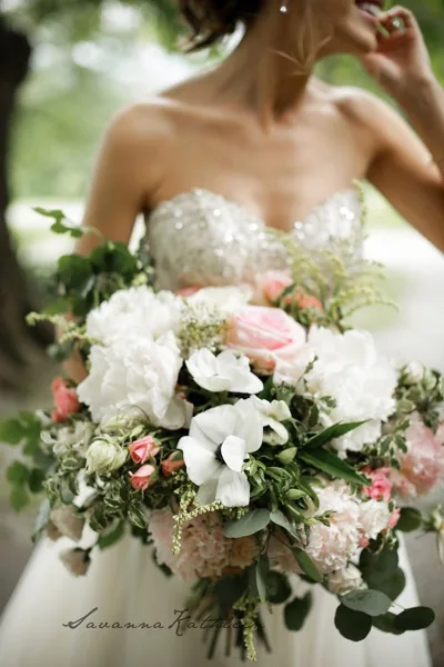A close-up of a bride holding a large, romantic bouquet of white and pink flowers with greenery.