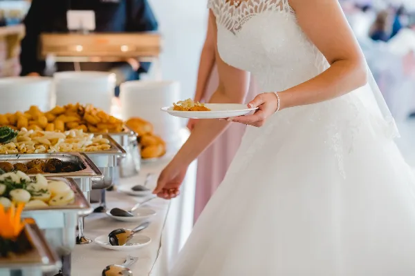 A bride in a white wedding dress serving food from a buffet line at a wedding event.