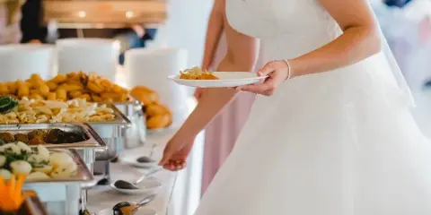 A bride in a white wedding dress serving food from a buffet line at a wedding event.