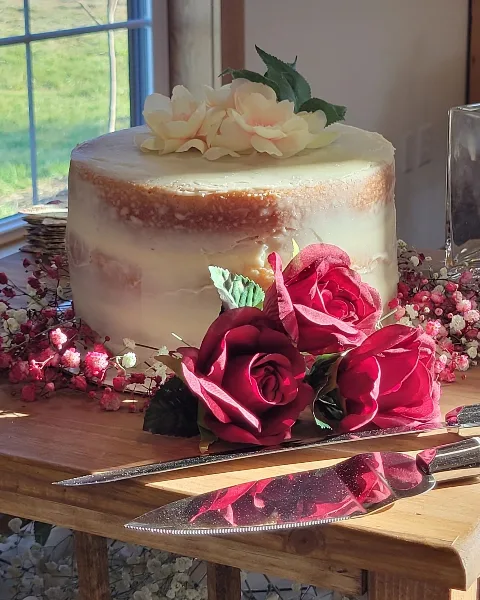 A semi-naked wedding cake topped with cream flowers and surrounded by red roses on a wooden table.