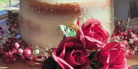A semi-naked wedding cake topped with cream flowers and surrounded by red roses on a wooden table.