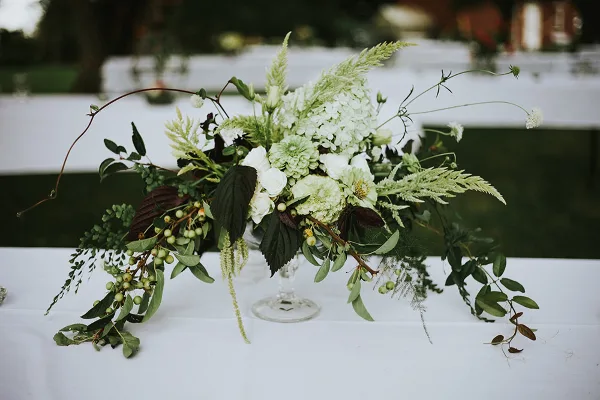 A lush floral centerpiece featuring white hydrangeas, greenery, and berries arranged in a glass vase on a white tablecloth.