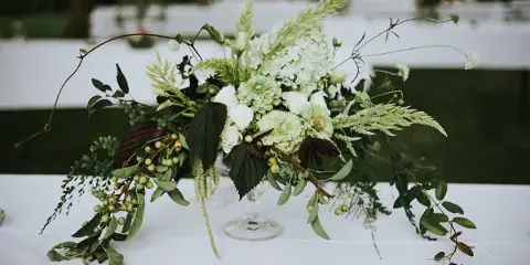 A lush floral centerpiece featuring white hydrangeas, greenery, and berries arranged in a glass vase on a white tablecloth.