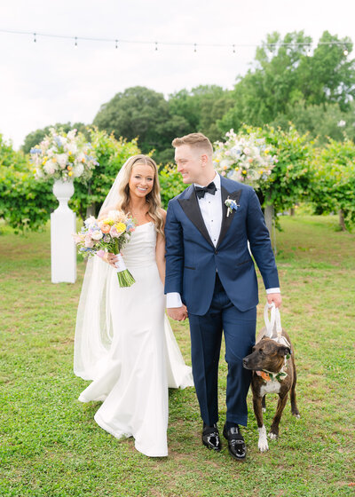 A bride and groom standing outdoors with a wedding bouquet and floral arrangements in the background.