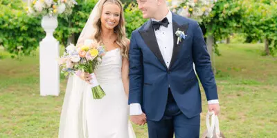 A bride and groom standing outdoors with a wedding bouquet and floral arrangements in the background.