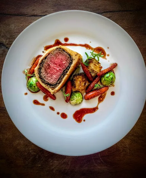 A plated Beef Wellington with medium-rare beef center, roasted carrots, Brussels sprouts, and a red wine reduction sauce on a white plate.