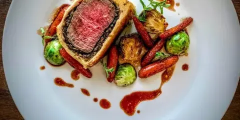 A plated Beef Wellington with medium-rare beef center, roasted carrots, Brussels sprouts, and a red wine reduction sauce on a white plate.