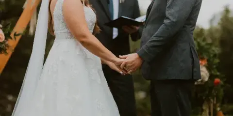A bride and groom holding hands during an outdoor wedding ceremony with an officiant in the background.