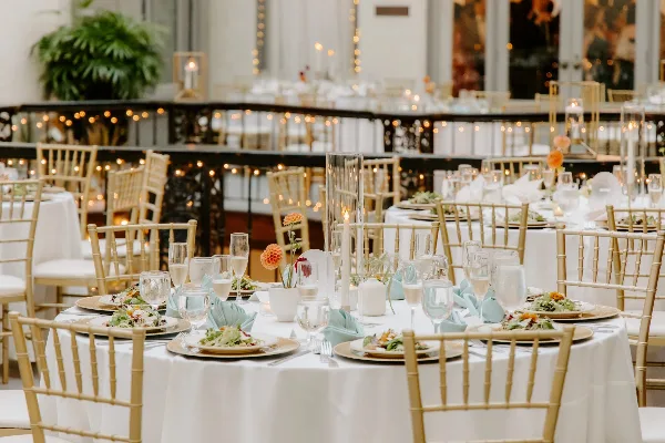 A wedding reception venue featuring round tables with white linens, gold chiavari chairs, and floral centerpieces.